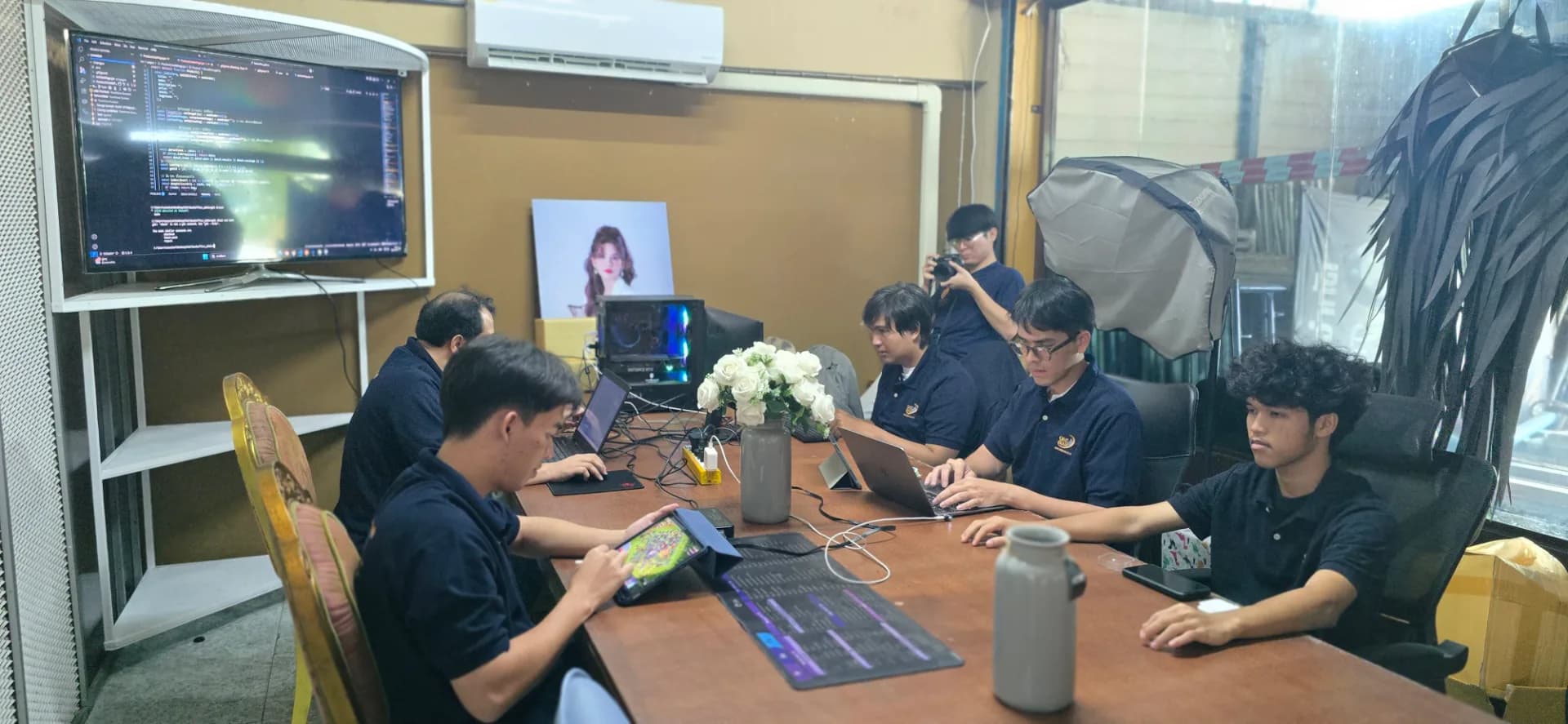 ภาพถ่ายบรรยากาศการทำงานจริง (Behind the Scenes) ของทีมงาน BizNewEra ในห้องทำงาน (Back Office) ที่เต็มไปด้วยความกระตือรือร้น โดยกลุ่มคนรุ่นใหม่ในทีมสวมเสื้อโปโลสีน้ำเงินเข้ม (Navy) ที่มีโลโก้บริษัท กำลังนั่งล้อมโต๊ะทำงานตัวยาวเพื่อปฏิบัติภารกิจต่าง ๆ ร่วมกันอย่างตั้งใจ ในภาพมีทีมงานฝ่ายต่าง ๆ ทั้งนักพัฒนาที่กำลังเช็กโค้ดบนหน้าจอใหญ่และโน้ตบุ๊ก, ฝ่ายคอนเทนต์และกลยุทธ์ที่กำลังทำงานผ่านแท็บเล็ตและอุปกรณ์ดิจิทัล, รวมถึงการจดบันทึกและเตรียมข้อมูล โดยรอบโต๊ะเต็มไปด้วยอุปกรณ์ทำงานอย่าง แล็ปท็อป สายเชื่อมต่อ ปลั๊กไฟ และแก้วน้ำ แสดงถึงการทำงานที่เป็นระบบและการร่วมแรงร่วมใจของทีมงานหลังบ้านในการขับเคลื่อนแบรนด์และพัฒนาธุรกิจในยุคดิจิทัล